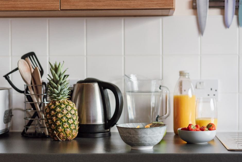 Kitchen table with whole pineapple and bowl of strawberries with bottle of juice and jar with water against wall covered with tile