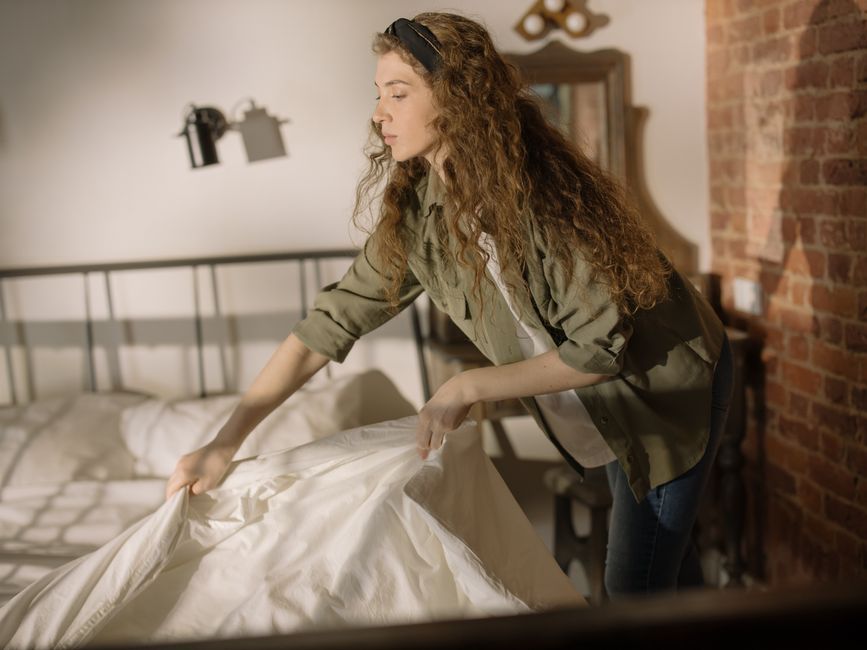 A young woman tidies up a cozy bedroom, emphasizing cleanliness and home comfort.