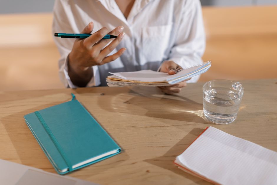 A person in a white shirt takes notes at a desk, holding a pen and notebook beside a glass of water.